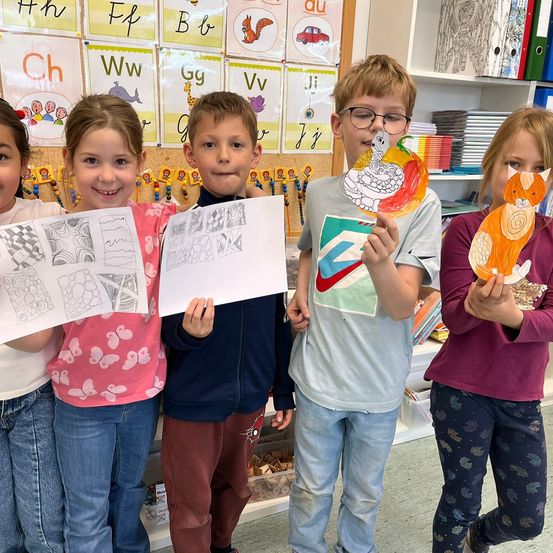 Fünf Kinder stehen in einem Klassenzimmer, halten Malblätter und Ausschnitte. Hinter ihnen sind Buchstaben und Bilder an der Wand.