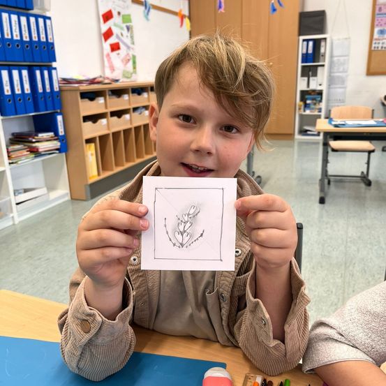 Ein junger Junge hält ein Blatt Papier mit einer Blumenskizze in der Hand, sitzt in einem Klassenzimmer, umgeben von Schreibtischen und Regalen.