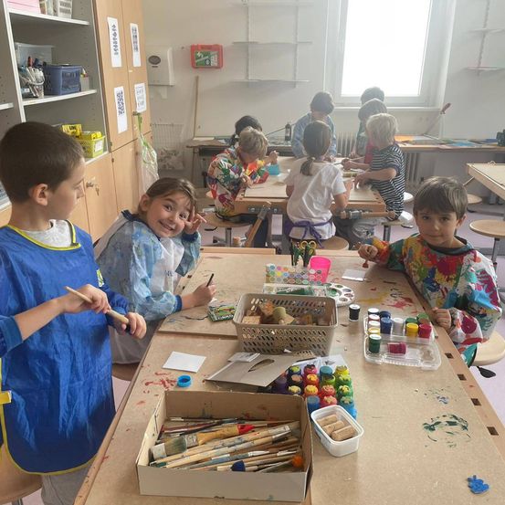 Kinder malen in einem Klassenzimmer. Einige sitzen an Schreibtischen, andere stehen. Kunstmaterialien wie Pinsel, Farben und Papier sind auf dem Tisch.