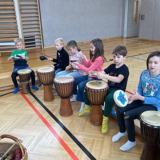 Eine Gruppe von Kindern sitzt in einer Reihe auf dem Boden und spielt Trommeln und andere Percussionsinstrumente. Sie befinden sich in einem Raum mit Holzboden und einer roten Linie auf dem Boden.