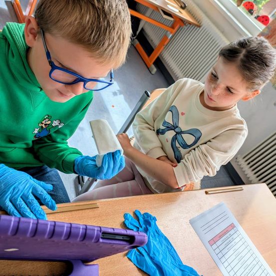 Zwei Kinder, die Handschuhe tragen, sitzen in einem Klassenzimmer. Ein Kind hält ein Papier, und beide scheinen an einer Aktivität beteiligt zu sein. Vor ihnen steht ein Schreibtisch mit einem Tablet und einem Papier. Hinter ihnen befindet sich ein Heizkörper und ein Fenster mit Blick auf eine Marienkäferfigur.