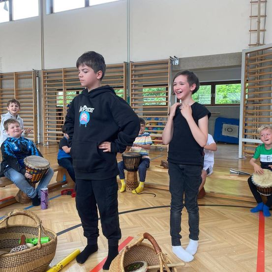 Kinder in einer Turnhalle, zwei stehen mit Trommeln. Einer lächelt, der andere hat die Hände auf dem Hosenbund. Dahinter sitzen andere Kinder auf einer Bank mit Trommeln.