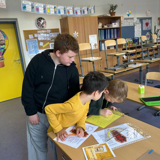 Ein Lehrer hilft zwei Schülern in einem Klassenzimmer, sie benutzen einen Winkelmesser auf einem Papierbogen. Das Klassenzimmer hat gelbe Türen, Holztische und verschiedene Poster.