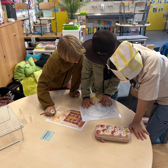 Drei Kinder arbeiten in einem Klassenzimmer an einem Projekt zusammen, eines benutzt einen Kompass und ein anderes einen Bleistift. Sie sind umgeben von verschiedenen pädagogischen Werkzeugen und Materialien.