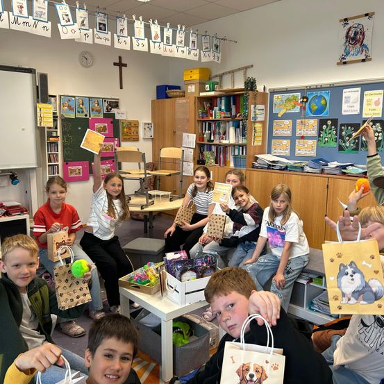 Eine Gruppe von Kindern in einem Klassenzimmer hält Geschenktüten. Sie sitzen um einen Tisch herum mit Spielzeug. Dahinter sind ein Bücherregal und Pinnwände mit verschiedenen Postern zu sehen.