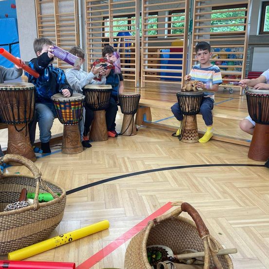 Kinder sitzen um Holztrommeln herum und spielen sie. Ein Junge trägt ein gestreiftes Hemd und gelbe Stiefel. Auf beiden Seiten befinden sich Körbe mit Musikinstrumenten.
