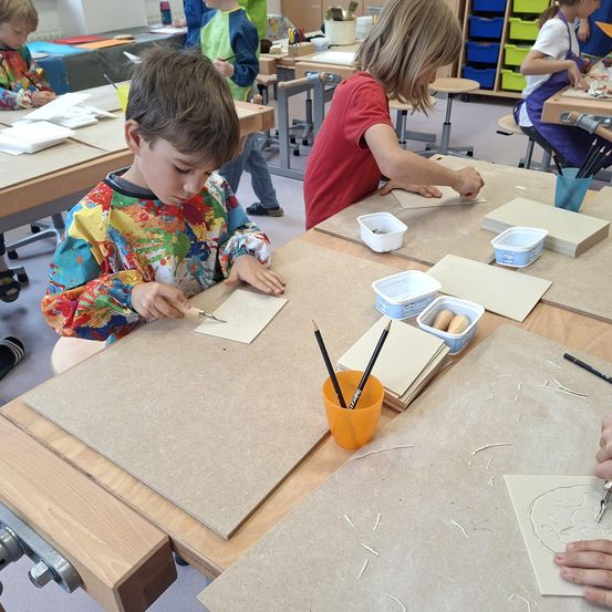 In einem Kunstunterricht sind Kinder mit Zeichnen und Basteln an ihren Tischen beschäftigt. Ein Junge benutzt ein Werkzeug zum Schneiden eines Stücks Papier.