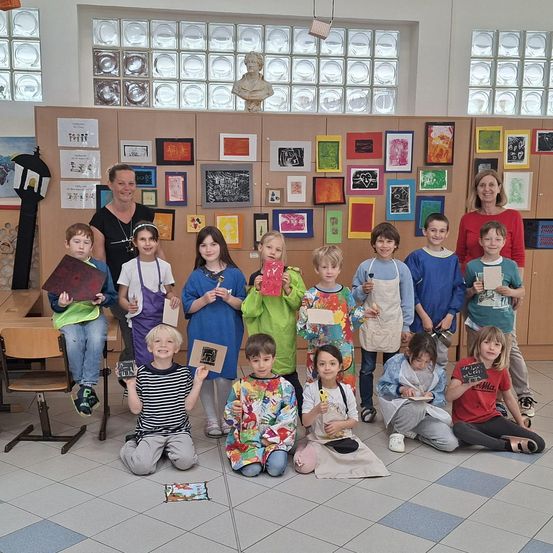 Eine Gruppe von Kindern in einem Klassenzimmer mit Kunst an den Wänden. Sie halten ihre Kunstwerke und sitzen in einer Reihe mit zwei Lehrern.