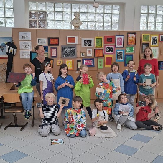 Gruppe von Kindern mit Malpinseln und Kunstwerken in einem Klassenzimmer, mit zwei Frauen hinter ihnen. Der Raum hat Kunstwerke an der Wand und eine Büste über den Frauen.