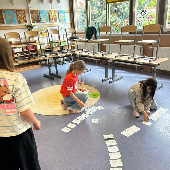 Drei Kinder sind in einem Klassenzimmer und ordnen Buchstaben auf dem Boden an. Ein Kind kniet, ein anderes sitzt und ein drittes steht in der Nähe. Im Hintergrund stehen leere Stühle und Tische.
