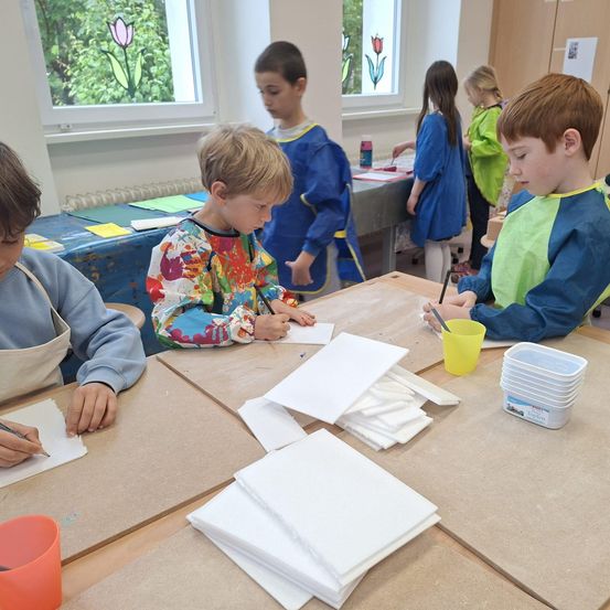 Kinder in einem Klassenzimmer, die Schürzen tragen, sind in einer künstlerischen Aktivität beschäftigt. Sie sitzen um einen Tisch herum, einer hält einen Pinsel, während andere auf Papier zeichnen. Der Raum hat große Fenster mit bunten Blumen.