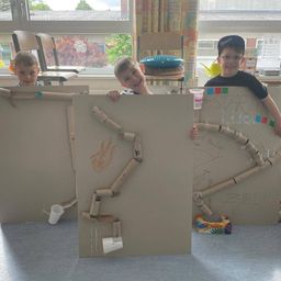Drei Jungen spielen mit einem Karton-Labyrinth mit einer Strecke aus Pappröhren, stehen hinter einer Pappwand, wobei einer einen Becher hält.