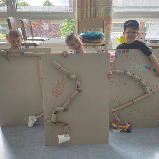 Drei Jungen spielen mit einem Karton-Labyrinth mit einer Strecke aus Pappröhren, stehen hinter einer Pappwand, wobei einer einen Becher hält.