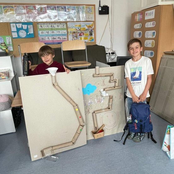 Zwei junge Jungen stehen hinter einer Pappkreation, die ein Rohrlabyrinth zeigt. Ein Junge lächelt und der andere hält eine Trichter. Sie befinden sich in einem Raum mit Stühlen und einem Schreibtisch.