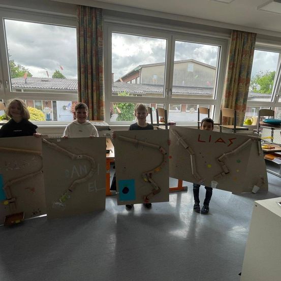 Vier Kinder posieren mit ihren Kunstprojekten in einem Raum mit großen Fenstern. Jedes Kind hält eine Kartonplatte mit einem einzigartigen Design. Draußen sind durch die Fenster Gebäude und Bäume zu sehen.