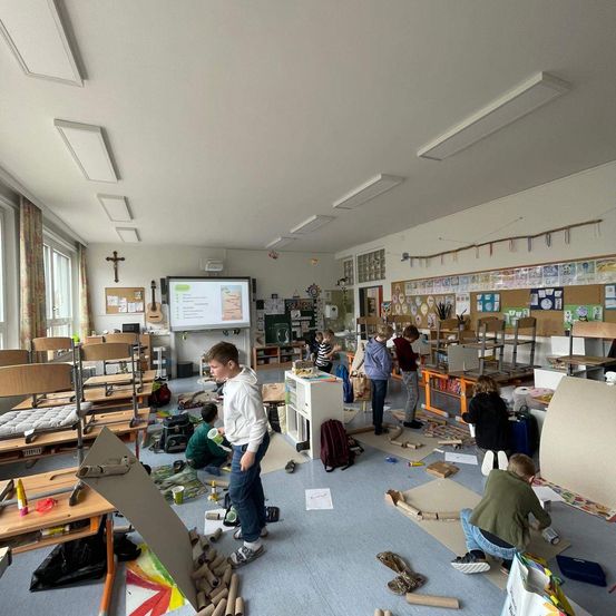 Ein Klassenzimmer mit einer Beamerleinwand zeigt eine Präsentation. Schüler arbeiten an Projekten mit Karton und anderen Materialien. Einige sitzen auf dem Boden, während andere stehen und arbeiten.