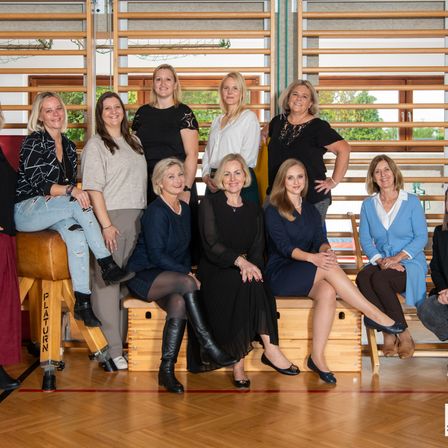 Eine Gruppe von Frauen posiert für ein Foto in einem Fitnessstudio. Sie sitzen auf Bänken und einige stehen dahinter. Der Holzboden und die Fitnessgeräte sind im Hintergrund.