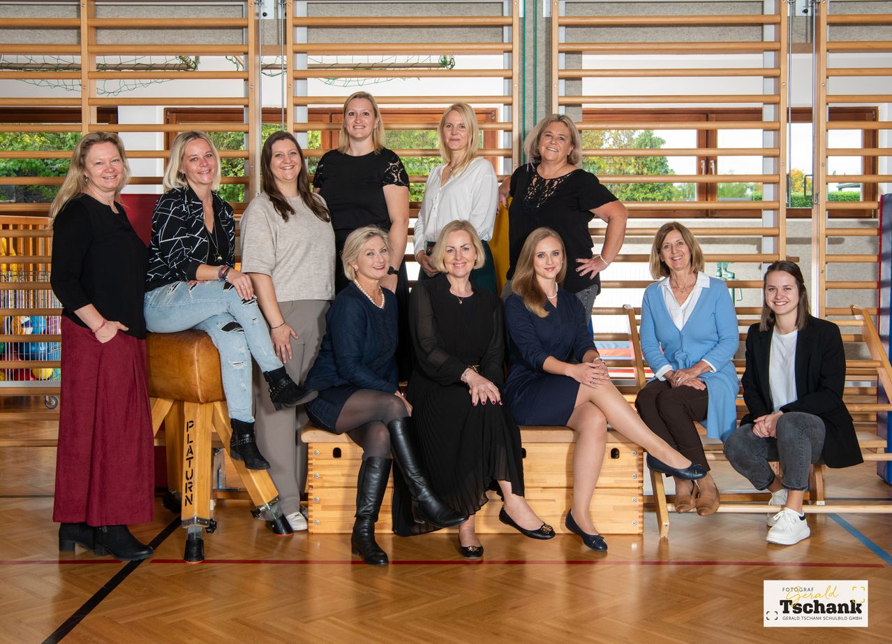 Eine Gruppe von Frauen posiert für ein Foto in einem Fitnessstudio. Sie sitzen auf Bänken und einige stehen dahinter. Der Holzboden und die Fitnessgeräte sind im Hintergrund.