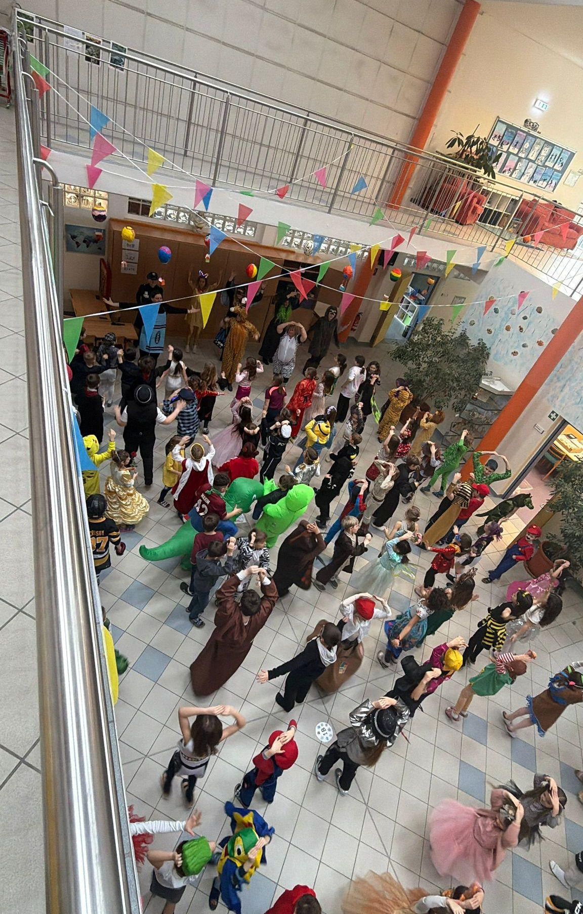 Eine große Gruppe von Kindern in verschiedenen Kostümen tanzt in einem dekorierten Saal, mit bunten Flaggen, die von der Decke hängen.