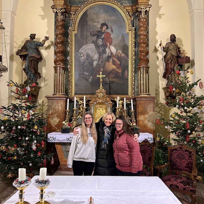 Drei Frauen stehen vor einem geschmückten Altar mit zwei Weihnachtsbäumen, die mit Ornamenten verziert sind. Ein Gemälde eines Ritters auf einem Pferd ist über dem Altar angebracht.