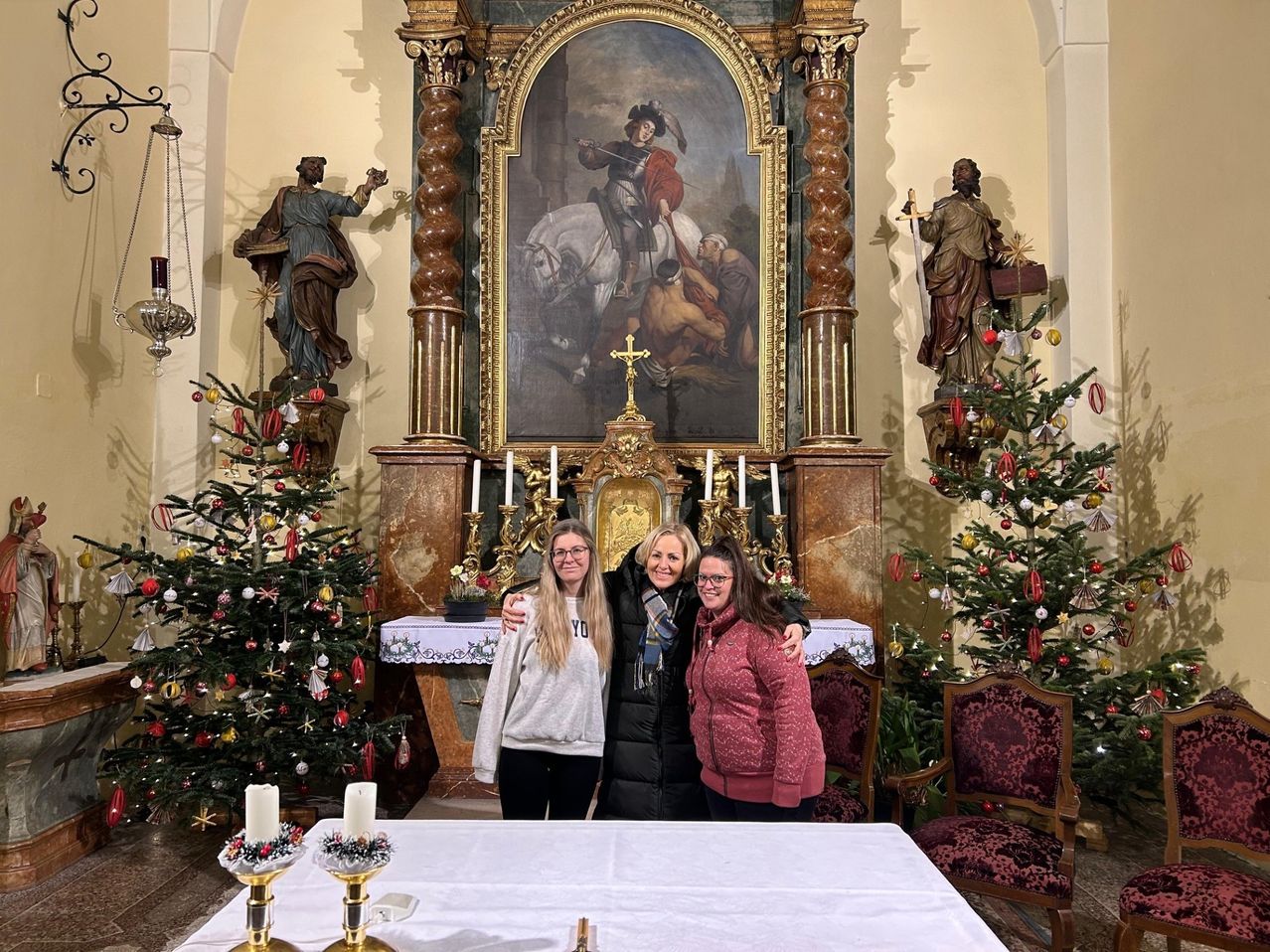 Drei Frauen stehen vor einem geschmückten Altar mit zwei Weihnachtsbäumen, die mit Ornamenten verziert sind. Ein Gemälde eines Ritters auf einem Pferd ist über dem Altar angebracht.