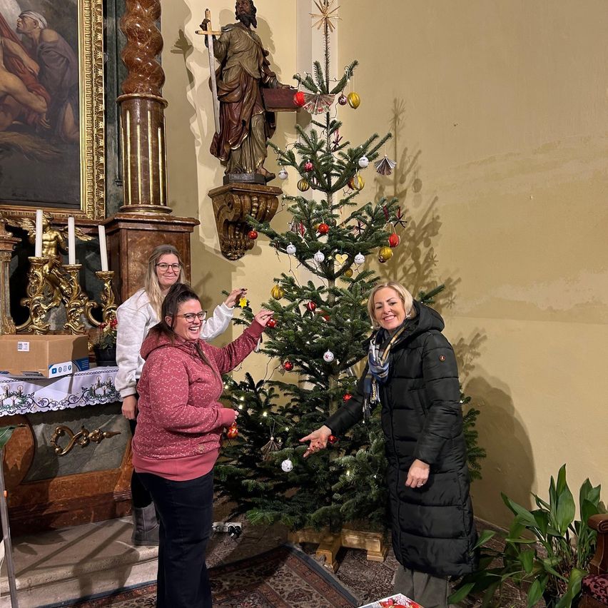Drei Frauen in Winterkleidung schmücken einen Weihnachtsbaum in einer Kirche. Eine Statue steht hinter dem Baum.