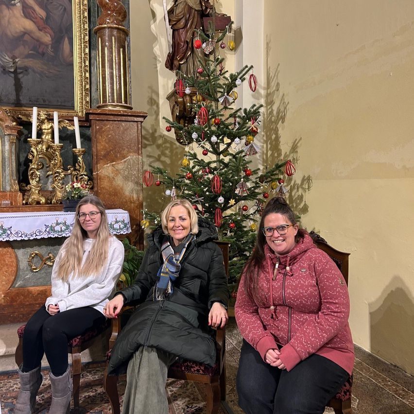 Drei Frauen sitzen in Stühlen vor einem geschmückten Weihnachtsbaum. Hinter ihnen ist eine Statue eines Heiligen zu sehen, und es gibt einen Marmoraltar mit einem weißen Tuch und zwei Kerzen.