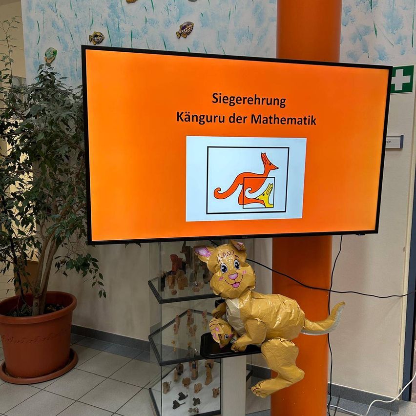 Ein Klassenzimmer-Display mit einem orangefarbenen Bildschirm zeigt ein Känguru-Motiv, mit einer Papier-Känguru-Dekoration auf einem nahen Regal.