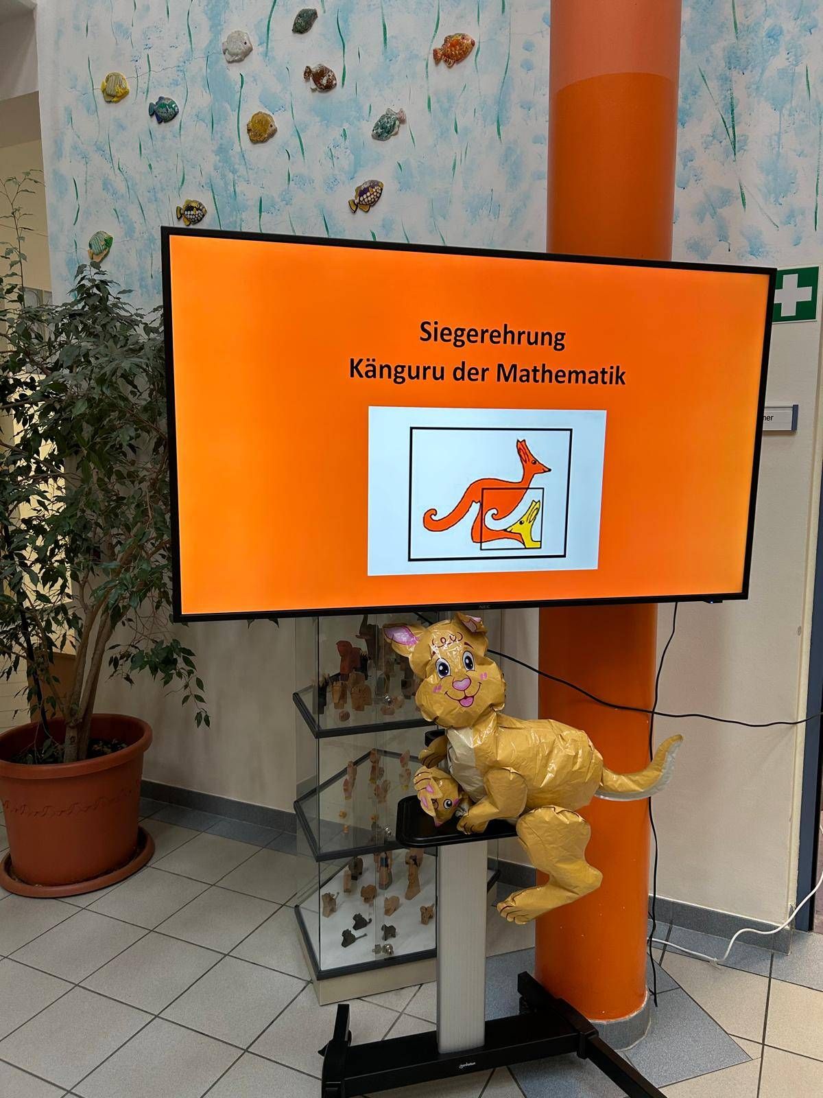 Ein Klassenzimmer-Display mit einem orangefarbenen Bildschirm zeigt ein Känguru-Motiv, mit einer Papier-Känguru-Dekoration auf einem nahen Regal.