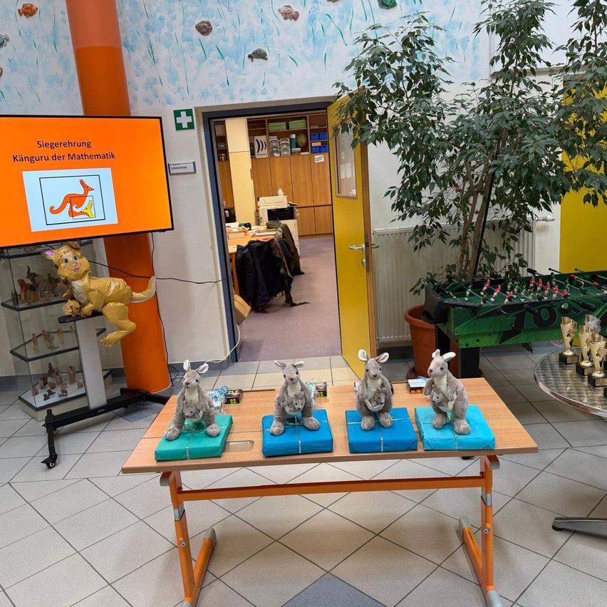 Vier Kängurufiguren sitzen auf einem Tisch mit blauem Geschenkpapier. Ein Monitor zeigt 'Sicherehrung Känguru der Mathematik'. Eine große Pflanze und eine Tür im Hintergrund.