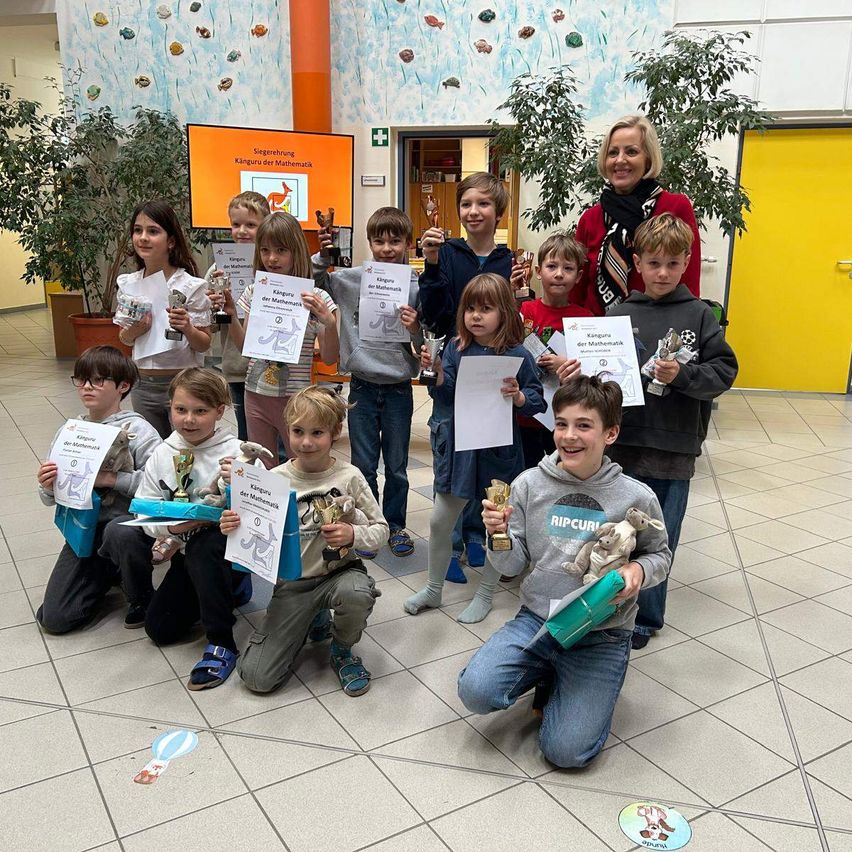 Kinder posieren für ein Foto mit Urkunden und Kuscheltieren. Eine Frau in Rot steht hinter ihnen. Der Ort ist drinnen, mit Pflanzen und einer Tür sichtbar.