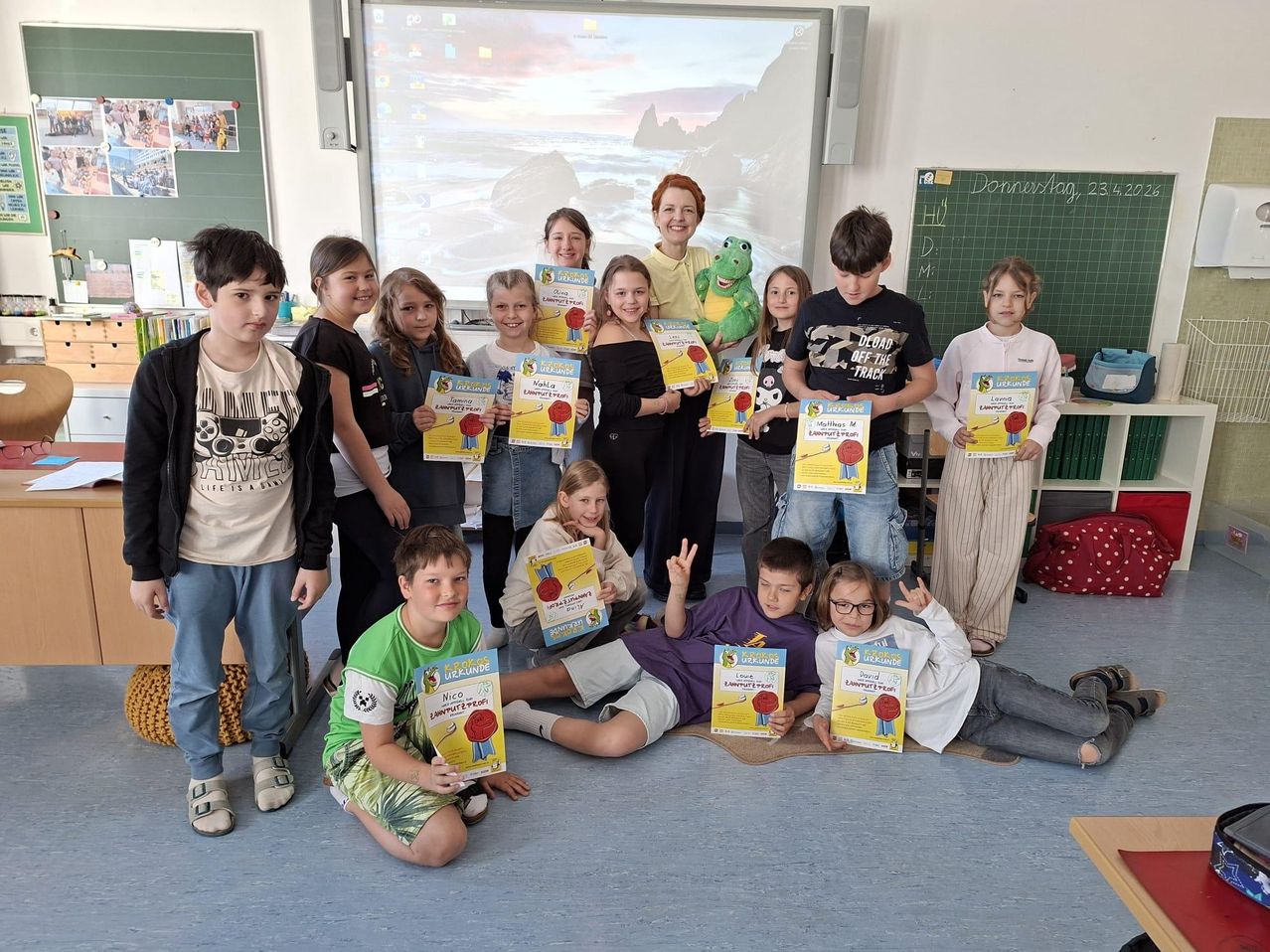 Kinder posieren für ein Foto im Klassenzimmer mit Zertifikaten und einer Lehrerin. Ein Froschspielzeug und ein Computerbildschirm sind im Hintergrund zu sehen.