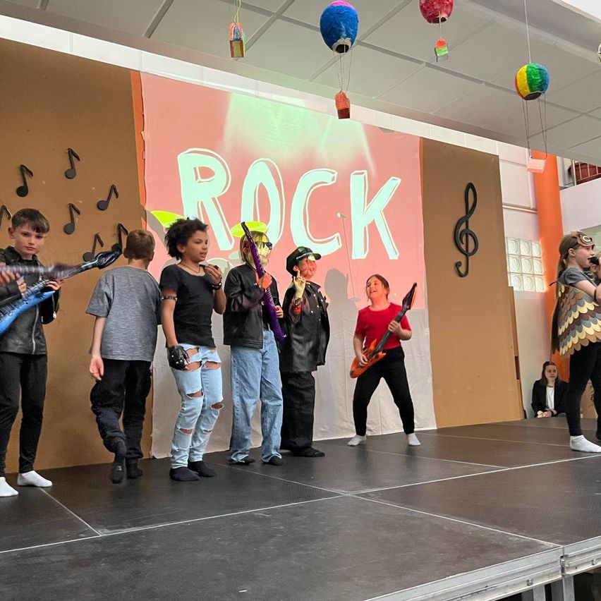 Eine Gruppe junger Leute steht mit Gitarren auf der Bühne. Eine Frau in einem roten Shirt spielt eine E-Gitarre. Der Hintergrund zeigt ROCK mit Musiknoten. Bunte Ballons hängen oben.