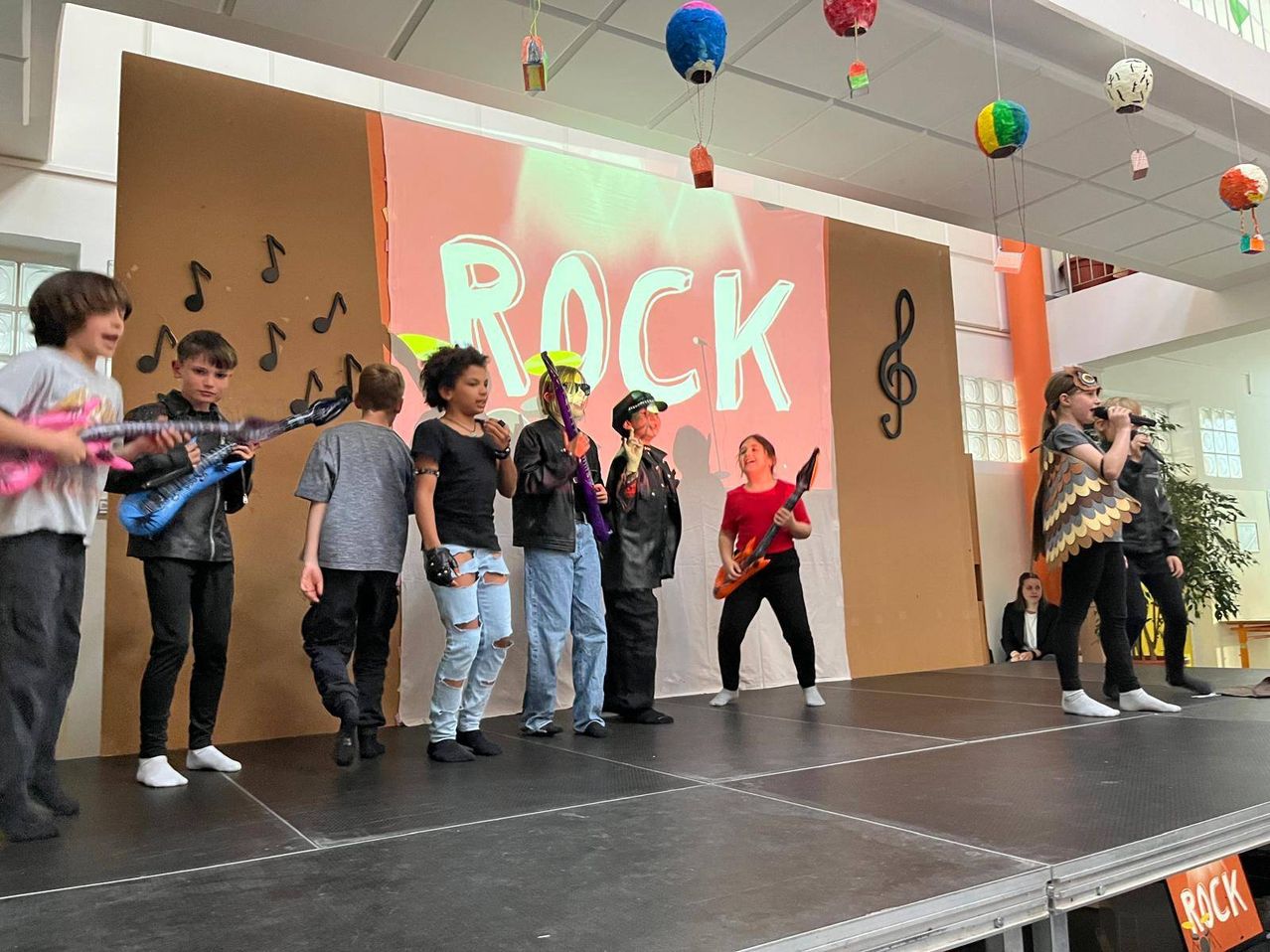 Eine Gruppe junger Leute steht mit Gitarren auf der Bühne. Eine Frau in einem roten Shirt spielt eine E-Gitarre. Der Hintergrund zeigt ROCK mit Musiknoten. Bunte Ballons hängen oben.