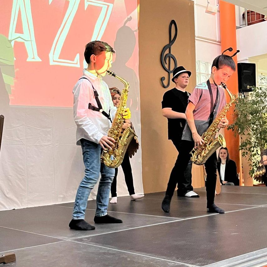 Zwei Jungen spielen Saxophon auf einer Bühne mit einem Hintergrund, auf dem 'Jazz' steht. Hinter ihnen beobachtet ein Junge. Ein Lautsprecher und eine musikalische Note sind an der Wand montiert.