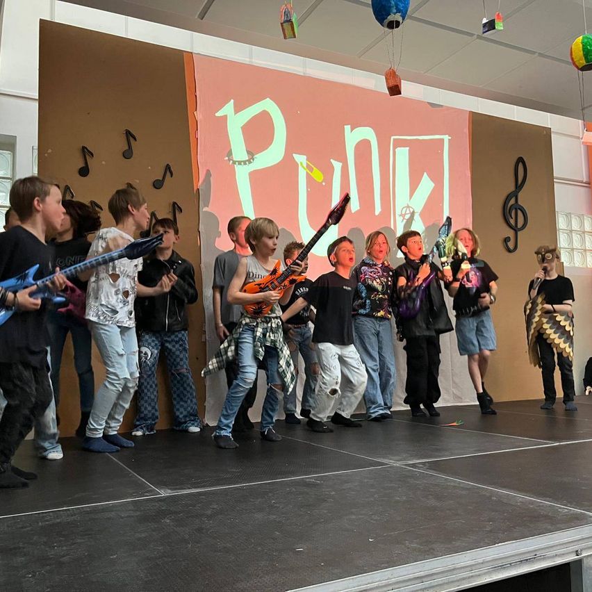 Eine Gruppe von Kindern auf der Bühne bei einer Punk-inspirierten Show, sie halten Gitarren und singen. Dahinter befindet sich ein Hintergrund mit dem Wort 'Punk' in großen Buchstaben.