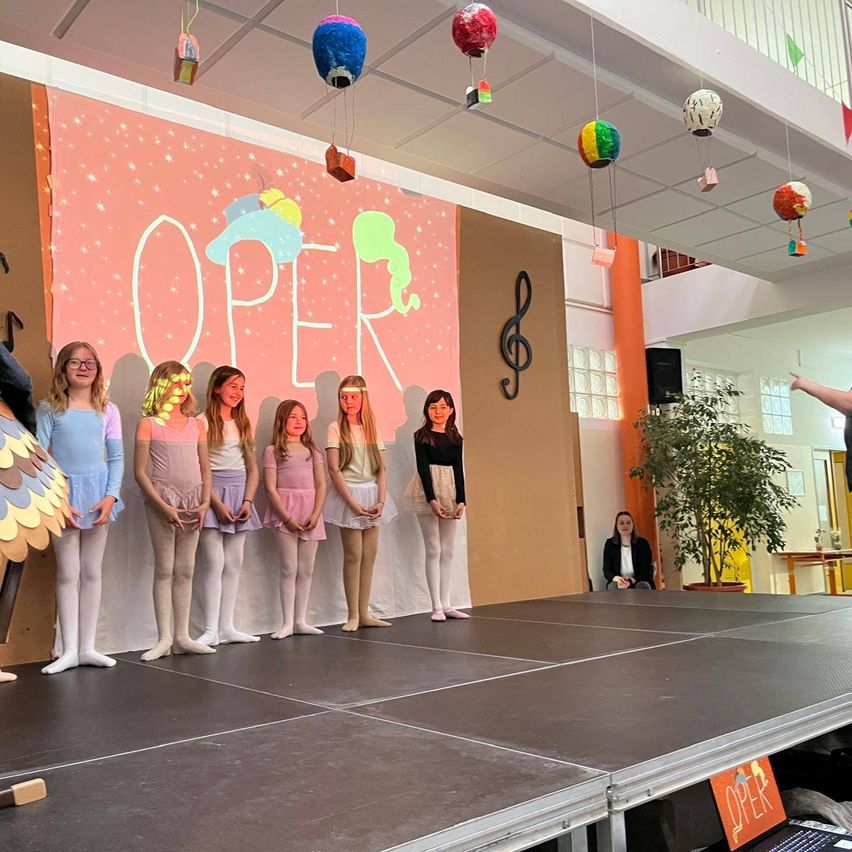 Eine Gruppe junger Mädchen in Ballettkleidung steht auf der Bühne vor einem Hintergrund mit der Aufschrift 'OPER' und Noten. Bunte Papierlaternen hängen von der Decke.