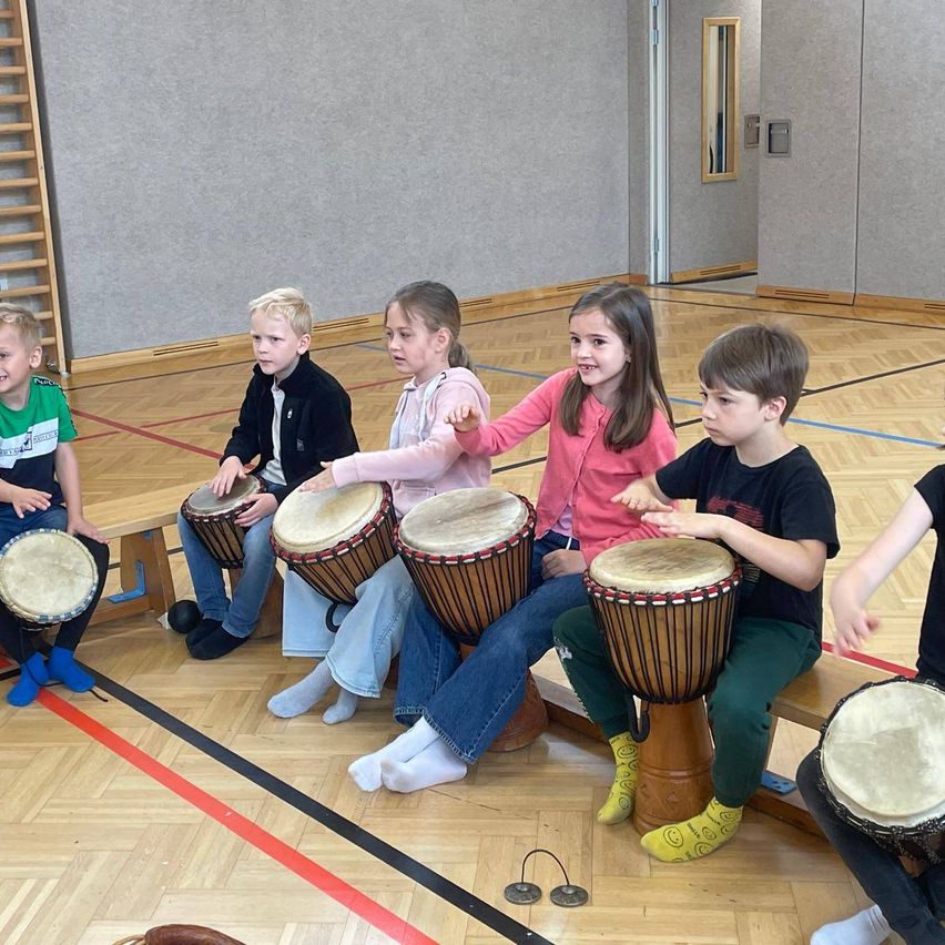 Mehrere Kinder sitzen auf einer Bank und spielen Trommeln. Sie lächeln und scheinen Spaß zu haben. Sie sind in einem Raum mit Holzboden.