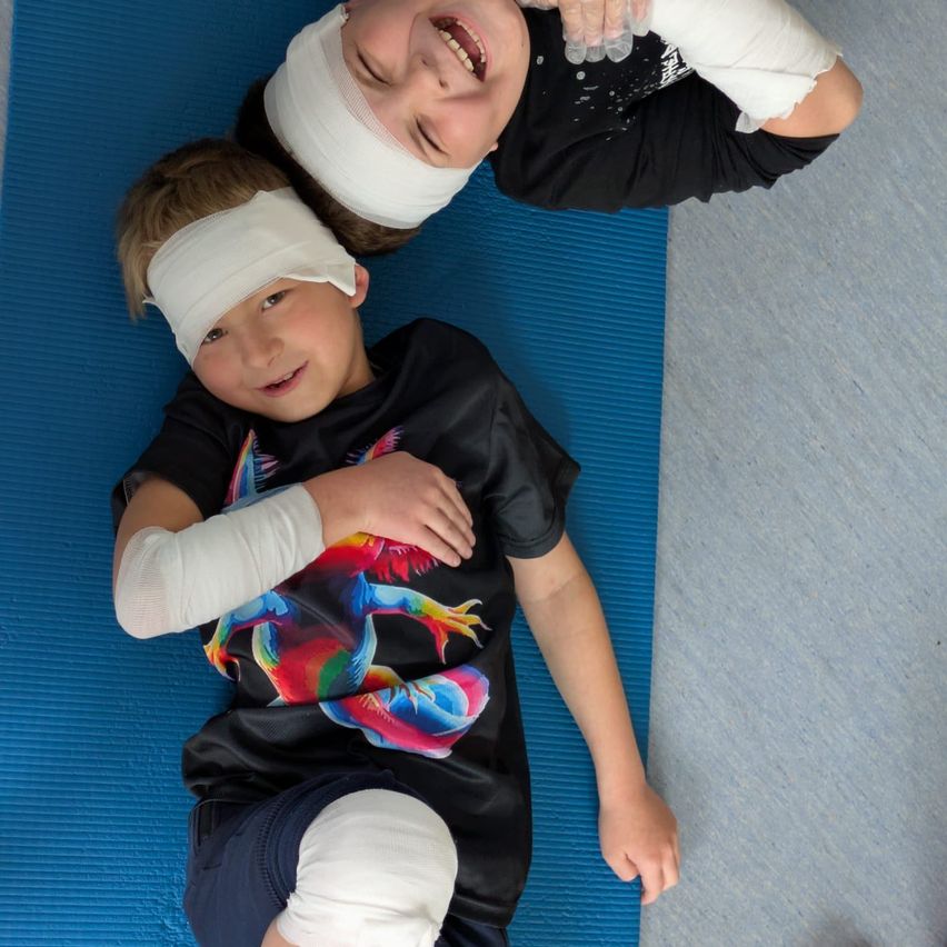 Zwei junge Jungen liegen auf einer blauen Yogamatte, beide haben Verbände an Kopf und Armen. Sie lächeln und scheinen für ein Foto zu posieren.