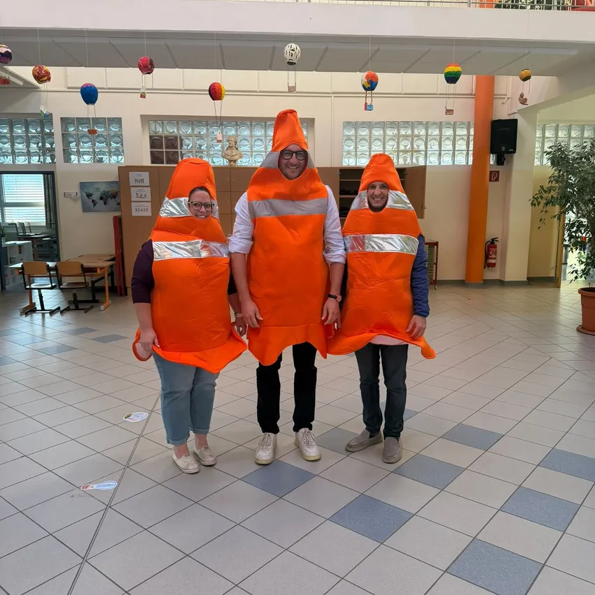 Drei Personen in orangefarbenen Verkehrskegel-Kostümen stehen in einem Flur. Die Person in der Mitte trägt eine Brille. Hinter ihnen ist ein Feuerlöscher an der Wand montiert.