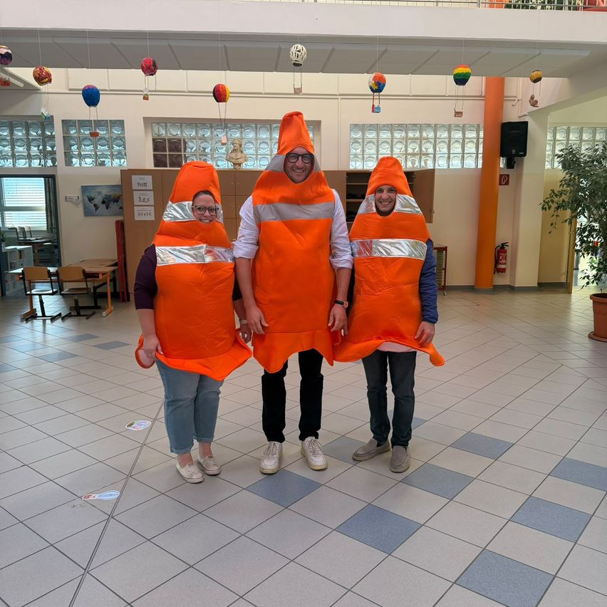 Drei Personen in orangefarbenen Verkehrskegel-Kostümen stehen in einem Flur. Die Person in der Mitte trägt eine Brille. Hinter ihnen ist ein Feuerlöscher an der Wand montiert.