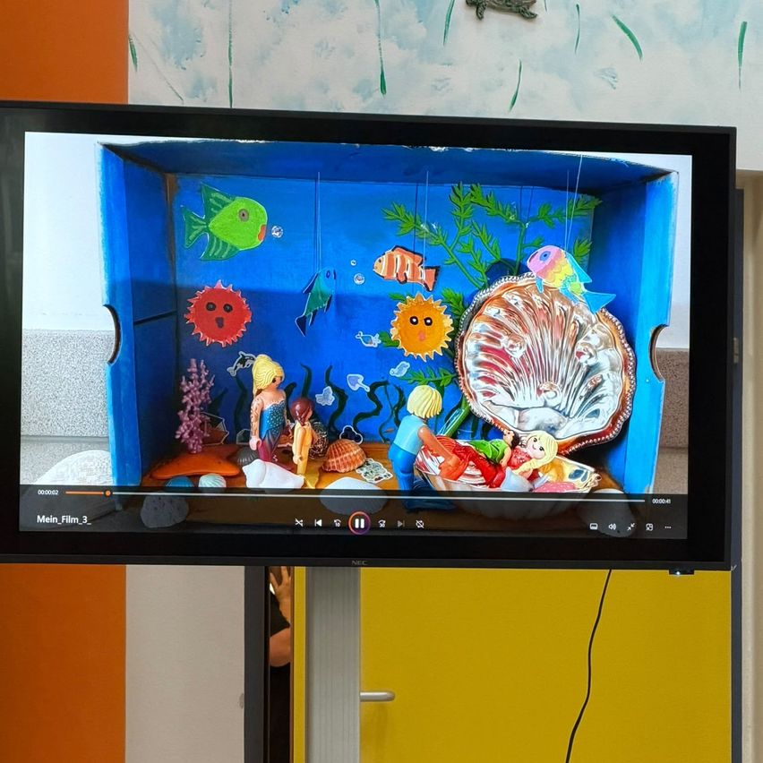 Ein Fernsehbildschirm zeigt ein farbenfrohes Aquarium mit verschiedenen Fischen, Meerestieren und einer Muschel. Das Aquarium besteht aus Pappe und hat einen blauen Hintergrund.