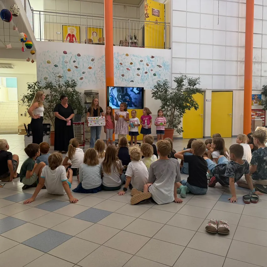 Eine Gruppe von Kindern sitzt im Kreis auf dem Boden und hört einer Präsentation zu. Zwei Frauen stehen vor ihnen, eine hält eine Box. Der Raum ist mit farbenfrohen Wandgemälden und Topfpflanzen dekoriert.
