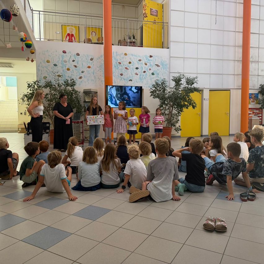 Eine Gruppe von Kindern sitzt im Kreis auf dem Boden und hört einer Präsentation zu. Zwei Frauen stehen vor ihnen, eine hält eine Box. Der Raum ist mit farbenfrohen Wandgemälden und Topfpflanzen dekoriert.