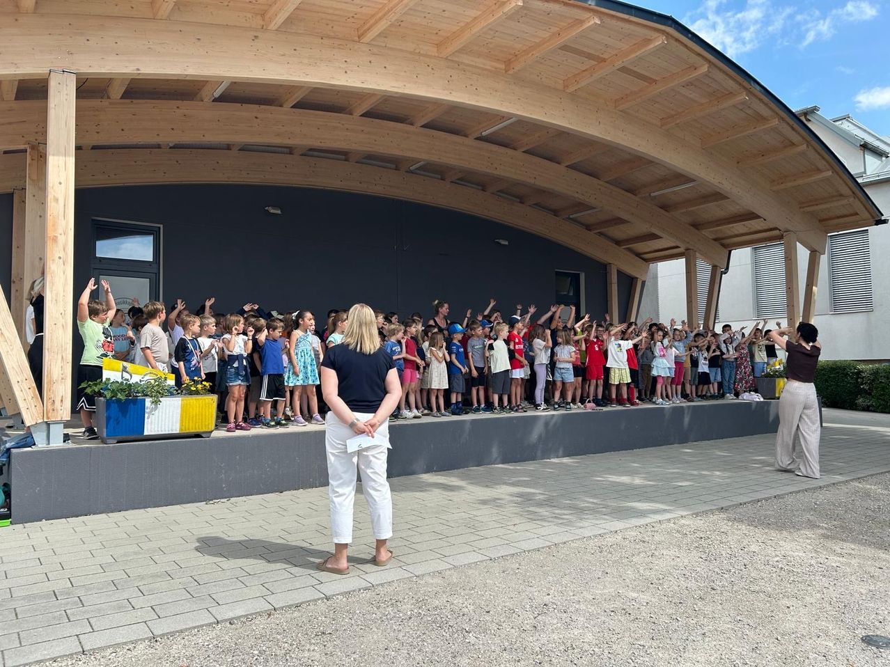 Eine Gruppe von Kindern steht auf einer Bühne mit einer Frau vor ihnen. Die Bühne ist von einem Holzdach umgeben. Die Kinder heben ihre Hände.