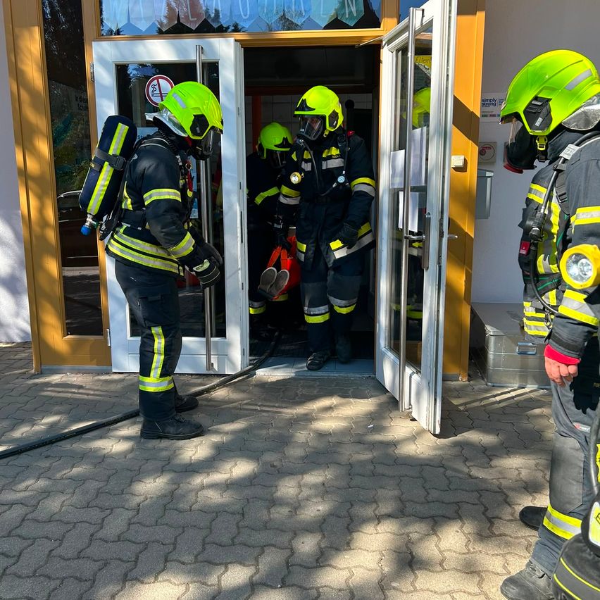 Feuerwehrleute in voller Montur verlassen ein Gebäude, einer trägt eine Trage. Das Gebäude hat eine Glastür und ein Schild am Fenster.