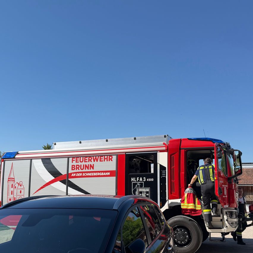 Ein Feuerwehrmann verlässt einen roten Feuerwehrwagen mit der Aufschrift 'Feuerwehr Brunn', geparkt neben einem schwarzen Auto. Die Szene ist unter einem klaren blauen Himmel.