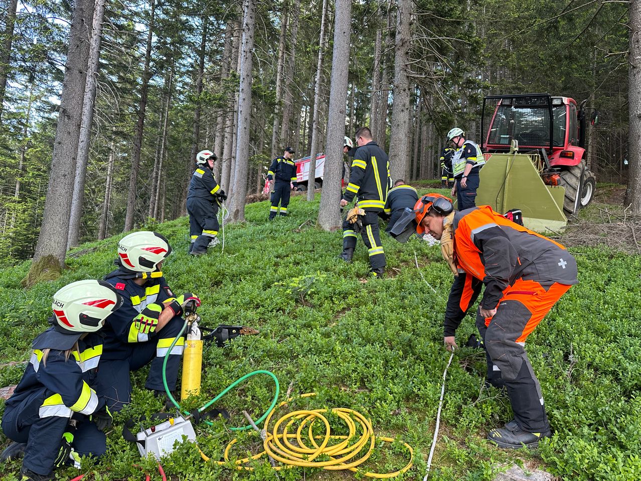 Feuerwehrleute üben eine Rettung im Wald, mit einem Feuerwehrauto und einem Zelt in der Nähe. Einige knien, andere stehen, alle tragen Helme und Handschuhe.