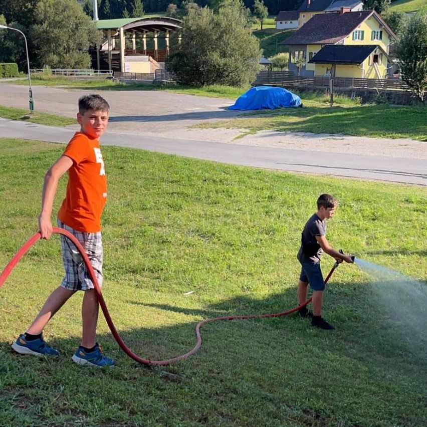 Zwei Jungen sprühen mit einem Gartenschlauch Wasser in einem Grasbereich. Im Hintergrund sind Häuser, ein Carport und ein überdachtes Auto zu sehen.