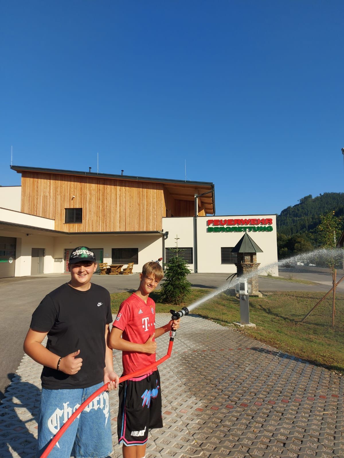 Zwei Jungen stehen vor einem Gebäude mit der Aufschrift 'Feuerwehr', halten einen Feuerwehrschlauch und sprühen Wasser. Ein Junge trägt ein rotes Shirt, der andere ein schwarzes. Der Himmel ist klar.